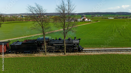 A Drone Side View of An Antique Steam Engine Traveling Along a Single RR Track, Thru Farmlands and Green Fields on a Spring Day
