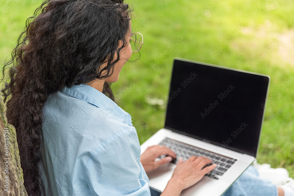 Young woman with laptop on the grass in the park sits by a tree. Remote work and training. Isolated on a black screen. Space for text. Girl with dark long curly hair typing on a computer Young woman with laptop on the grass in the park sits by a tree. Remote work and training. Isolated on a black screen. Space for text. Girl with dark long curly hair typing on a computer