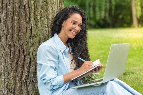 Portrait of a lovely stylish positive Brazilian or Hispanic female student, sitting on the grass near the tree, with a laptop and notepad, doing homework, taking notes, smile at camera