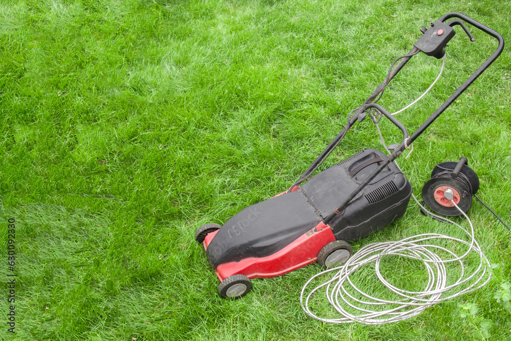 red electric lawn mower on a green lawn with wires and an electric extension cord, portable socket red electric lawn mower on a green lawn with wires and an electric extension cord, portable socket
