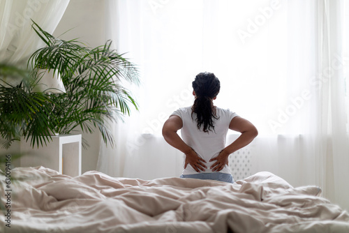 Backache Concept. Young Black Woman Massaging Aching Back While Sitting In Bed