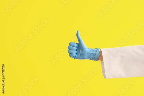Female doctor's hand in medical glove showing thumb-up on yellow background