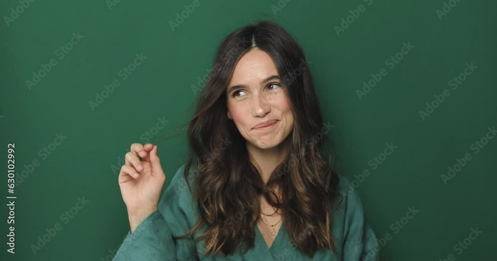 Beautiful young woman in green blouses winds a curl on a finger posing isolated over a green background in studio. Lovely coquettish brunette woman, winking playfully, flirting.
