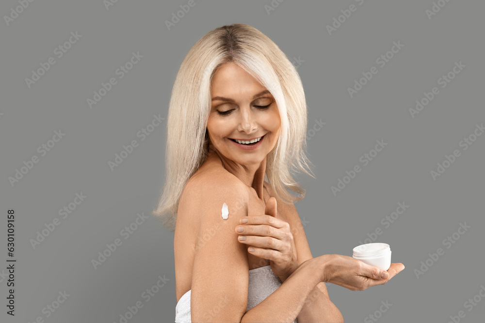 Beautiful Mature Blonde Woman Applying Moisturising Cream On Shoulder Over Grey Background Beautiful Mature Blonde Woman Applying Moisturising Cream On Shoulder Over Grey Background