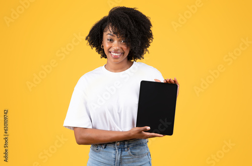 Cheerful young black curly lady in white t-shirt shows tablet with blank screen, isolated on orange background
