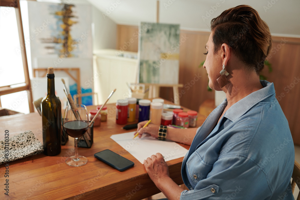 Mature woman drinking wine when drawing pictures at desk in her studio Mature woman drinking wine when drawing pictures at desk in her studio