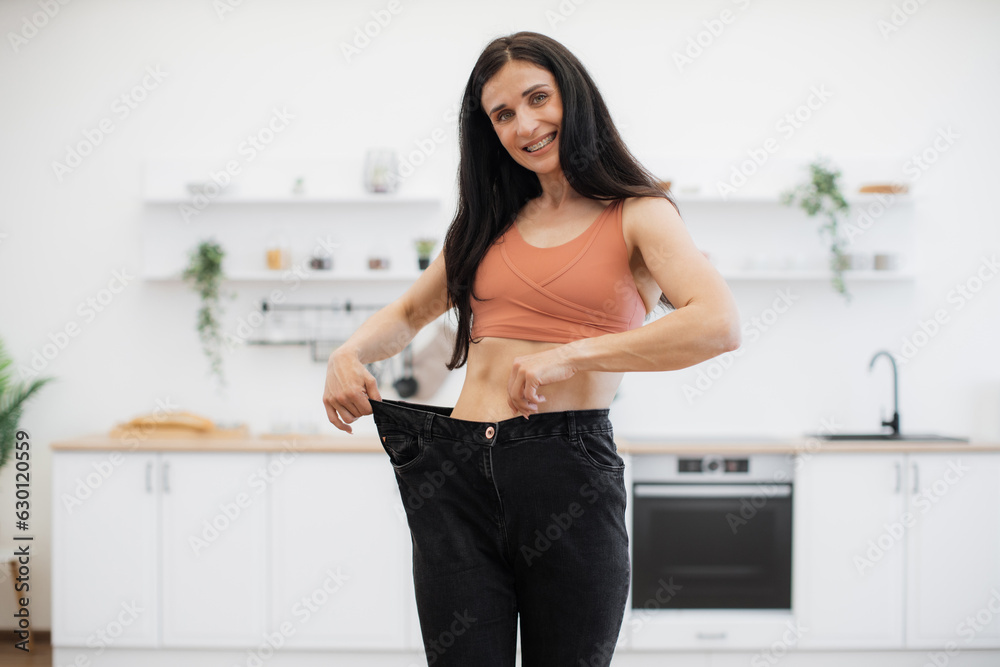 Surprised lady in terracotta top looking at black oversized jeans on thin waist while standing in kitchen of apartment. Sporty slim woman enjoying amazing achievements of well-balanced diet at home. Surprised lady in terracotta top looking at black oversized jeans on thin waist while standing in kitchen of apartment. Sporty slim woman enjoying amazing achievements of well-balanced diet at home.