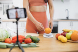 Close up view of slender woman weighing orange on smart digital scale while gadget on tripod shooting the process. Lady in good shape giving advice on improving diet with low-calorie products.