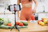 Focus on female in activewear checking nutrition value of sweet pepper via scales in home kitchen. Sports-loving food influencer filming dietary advice about benefits of organic products on phone.