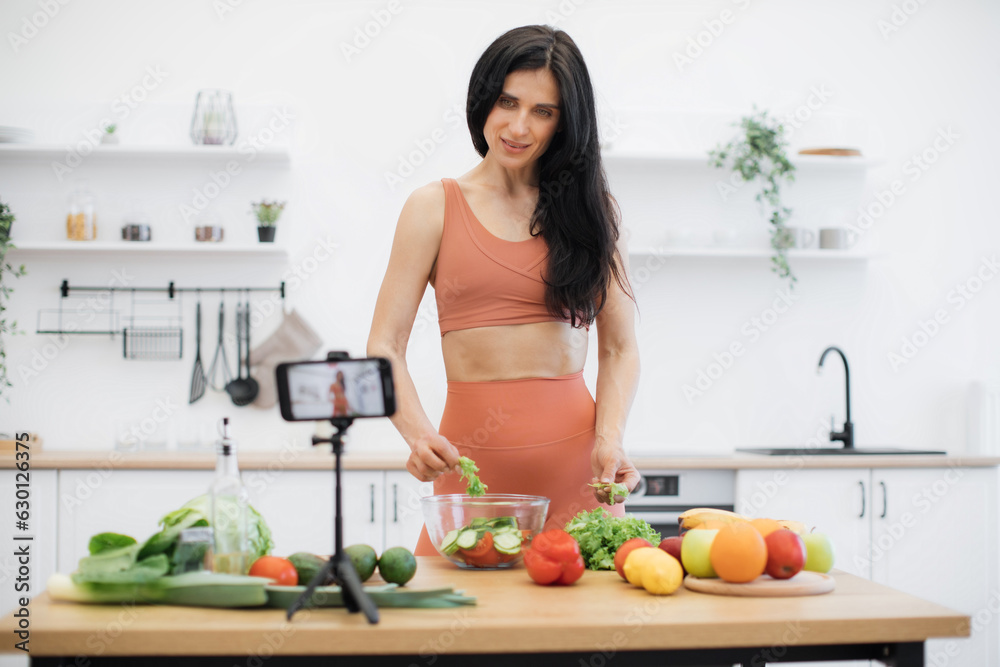 Cheerful caucasian adult adding leafy greens to salad bowl while looking at camera of cell phone in kitchen. Efficient food blogger starting online cooking course about healthy eating habits at home.
