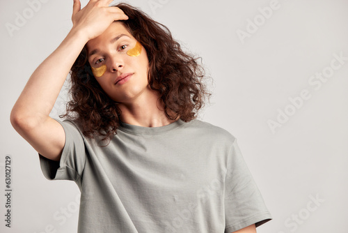 Young handsome long-haired guy applying under eye hydrogel patches on his face. Caucasian brown-haired millennial man practices skin care routine to keep healthy and youthful looking. Studio portrait.