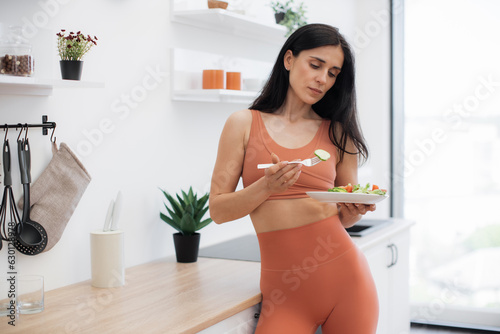 Billede på lærred Pensive fitness person dressed in gym clothes standing in kitchen and looking at plate with greens