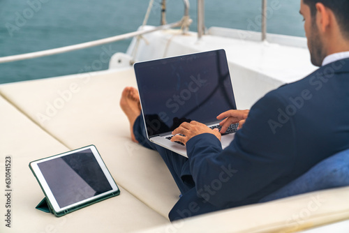 Confidence businessman working on laptop computer and talking on mobile phone for global corporate business while travel on luxury private catamaran boat yacht sailing in the ocean on summer vacation. © CandyRetriever 