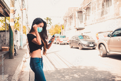 A slim Filipina woman in a crop top uses a ride sharing app and waits for a car while wearing a face mask to shield from pollution. In a typical Manila neighnorhood. photo