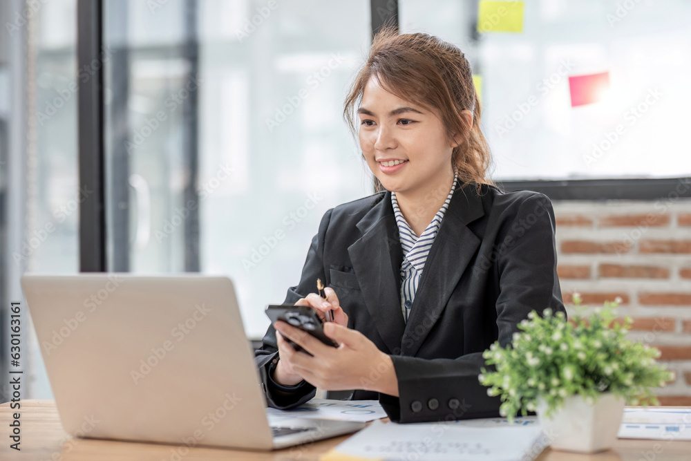 Asian businesswoman using smartphone to do accounting work with laptop and graph of financial reports in office