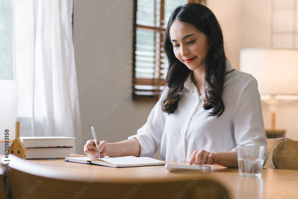 A millennial Asian businesswoman is sitting and working with laptop computer in her home kitchen, while also enjoying a relaxing cup of coffee.