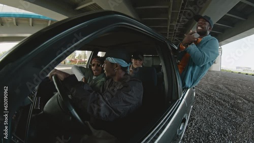 Four young diverse men wearing oversized clothes, bandanas and caps listening and dancing to hip hop and rap music in black retro car with open windows photo