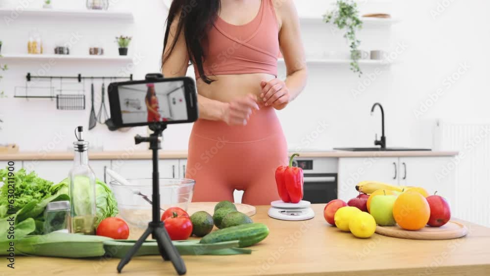 Focus on female in activewear checking nutrition value of sweet pepper via scales in home kitchen. Sports-loving food influencer filming dietary advice about benefits of organic products on phone.