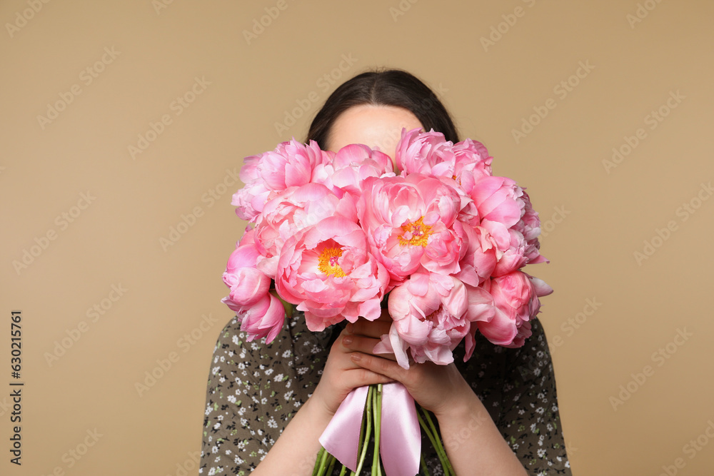 Young woman covering her face with bouquet of peonies on light brown background