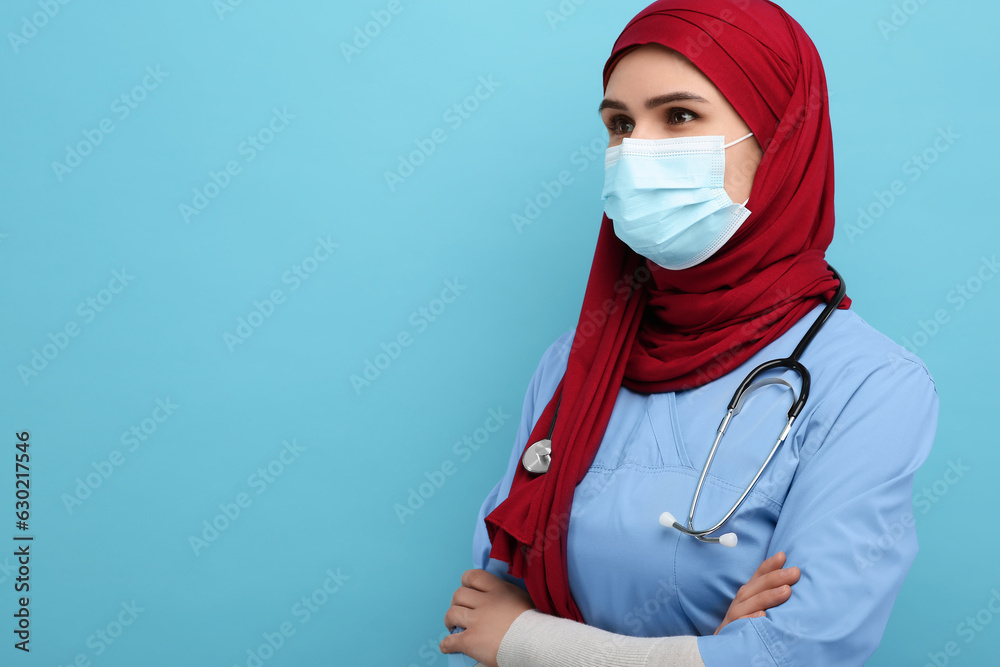 Muslim woman wearing hijab, medical uniform and protective mask on light blue background, space for text Muslim woman wearing hijab, medical uniform and protective mask on light blue background, space for text