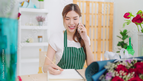 Beautiful young asian women owner floral store in apron talking on smartphone taking order with client and writing on notebook. Small business concept