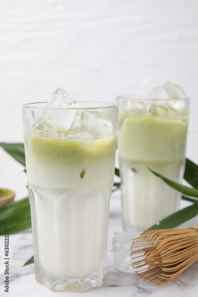 Glasses of tasty iced matcha latte, bamboo whisk and leaves on white marble table