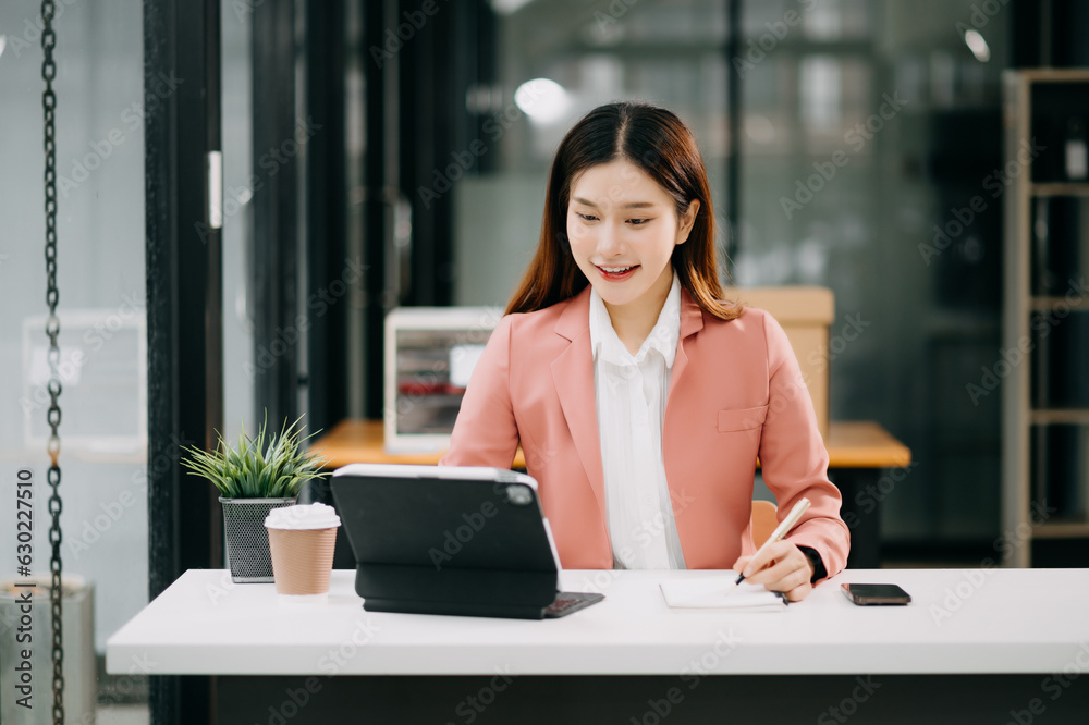 Young beautiful Asian woman typing on tablet and laptop while sitting at the working white table modern office Young beautiful Asian woman typing on tablet and laptop while sitting at the working white table modern office