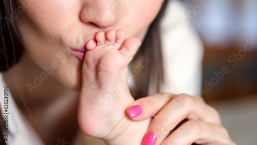 Mother kisses baby daughter feet playing on bed in bedroom. Infant girl kicks feet enjoying attention and careness from loving parent