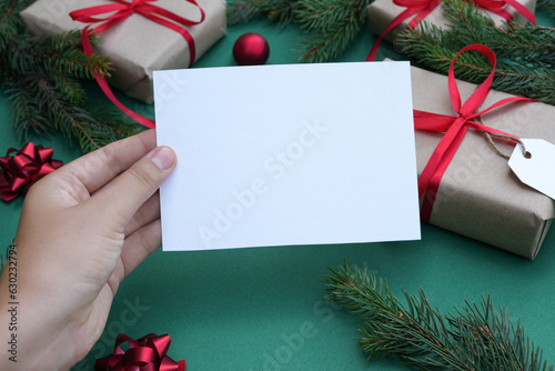 Blank paperin female hands. Flat lay of green background with gift boxes, Christmas decoration and fir tree branches. Top view mock up and copy space for text. Christmas background.