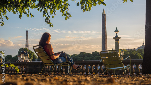 Jeune femme assise sur un chaise publique du Jardin des Tuileries lit un livre au soleil, vue sur Tour Eiffel et l'Obélisque de la place de la Concorde