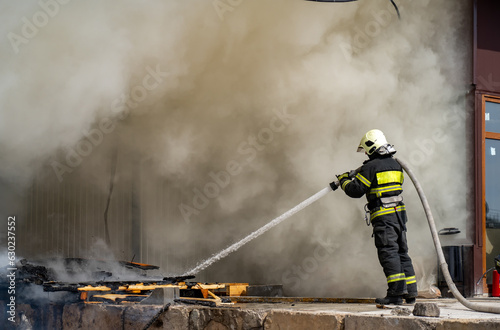 Amidst thick smoke, an unidentified firefighter unleashes a forceful spray of water from a hose, targeting the doorway of a building.