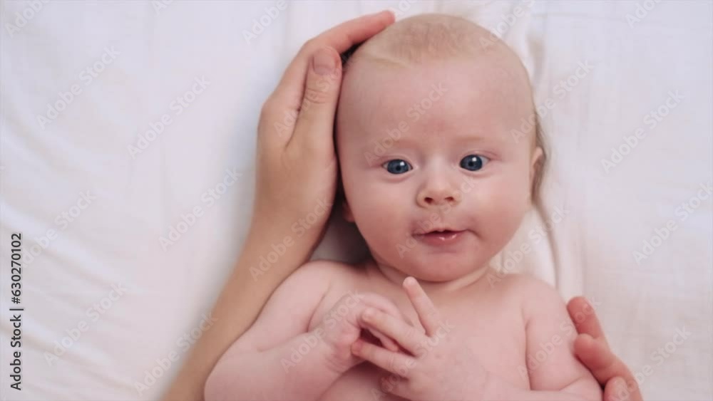 Close-up portrait of smiling adorable newborn baby lying on cozy light bed at home, loving young mother carefully touches cheeks and strokes head cute infant son. Happy childhood moments concept Close-up portrait of smiling adorable newborn baby lying on cozy light bed at home, loving young mother carefully touches cheeks and strokes head cute infant son. Happy childhood moments concept