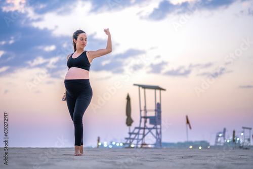 Nine months pregnant woman in sportswear showing muscles, standing on the sandy beach at sunset. Healthy lifestyle, sport and pregnancy concept.