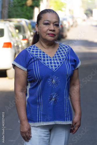 Mature Mexican lady looking at the camera smiling,wearing a traditional mexican embroidered blouse standing, with out of focus background of street. Latin woman looking at camera
