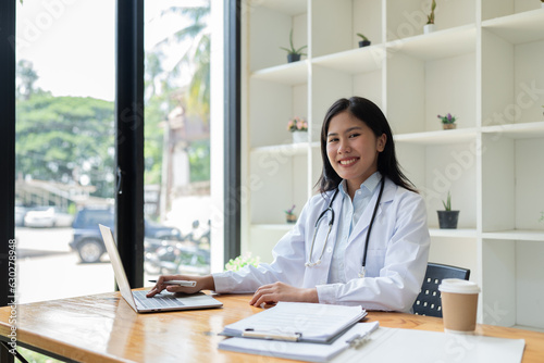 Asian female doctor sits at a desk working in the office, smiling and looking at the camera.
