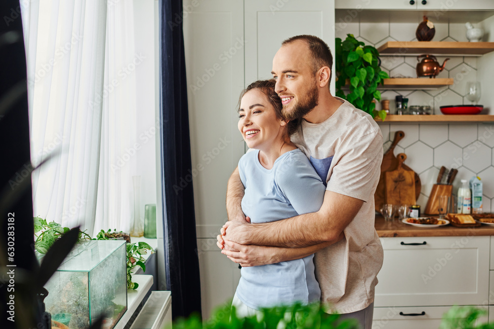 Positive man in homewear hugging brunette girlfriend while standing near plants and window at home Positive man in homewear hugging brunette girlfriend while standing near plants and window at home