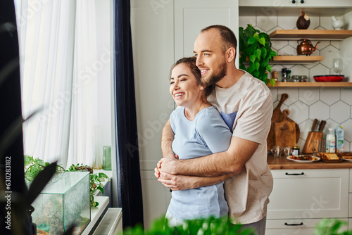 Positive man in homewear hugging brunette girlfriend while standing near plants and window at home