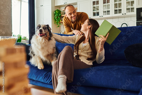Smiling couple spending time with book and border collie dog on couch near popcorn in living room