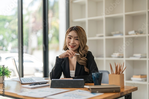 Portrait of young attractive asian woman with relaxing while sitting at her office desk, smiling and looking at camera