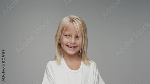 Portrait of Young Girl with Smiling Face Looking at Camera in Colour Studio Shot. Happy Cheerful Kid Isolated Alone on Grey Background Closeup. Emotional Child with Bright Blue Eyes in White Clothes