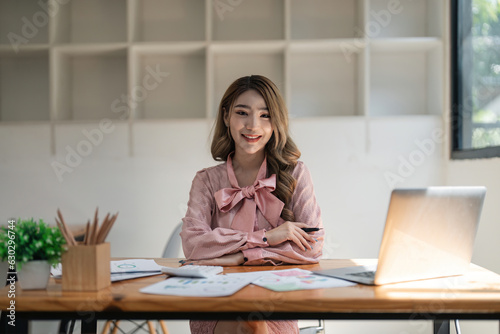 Portrait of young attractive asian woman with relaxing while sitting at her office desk, smiling and looking at camera