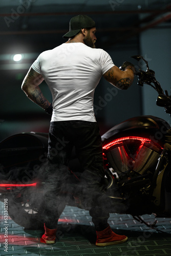 Athletic bearded biker man posing with motorbike with his back to the frame © Vitalii But
