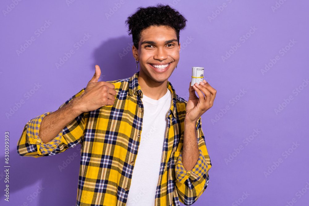 Photo of cheerful nice person toothy smile hand hold roll dollar bills demonstrate thumb up isolated on violet color background Photo of cheerful nice person toothy smile hand hold roll dollar bills demonstrate thumb up isolated on violet color background