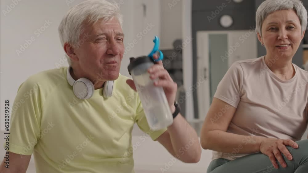 Tilt up shot of cheerful senior man with headphones sitting next to his wife in gym, drinking water and talking with trainer while resting after group workout Tilt up shot of cheerful senior man with headphones sitting next to his wife in gym, drinking water and talking with trainer while resting after group workout