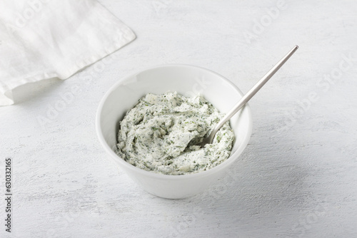 White bowl with cottage cheese cream mixed with herbs and garlic on a light gray background. Cooking appetizer or toppings for stuffing vegetables, vegetarian food