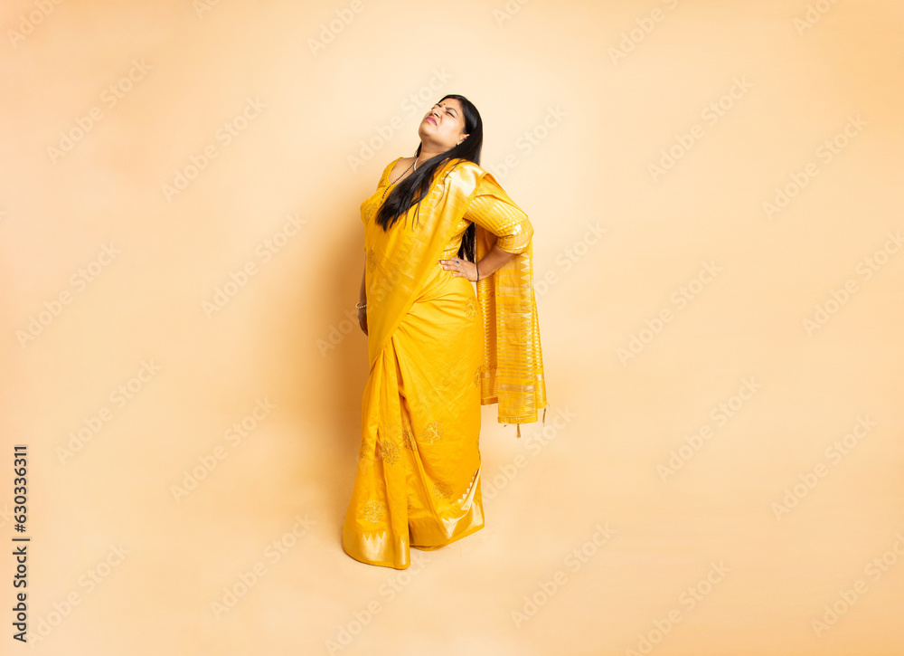 Indian woman having back pain standing isolated on beige studio background, Low waist hurt massage, Female wearing sari suffering from backache Indian woman having back pain standing isolated on beige studio background, Low waist hurt massage, Female wearing sari suffering from backache