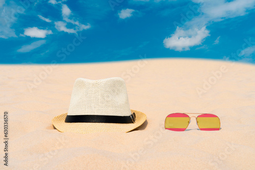Sunglasses with glasses in the form of the flag of Spain and a hat lie on the sand against the blue sky. The concept of summer holidays, travel and tourism in Spain