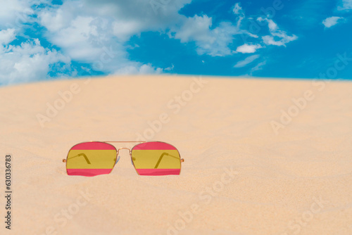 Sunglasses with glasses in the form of the flag of Spain lie on the sand against the blue sky. The concept of summer holidays, travel and tourism in Spain