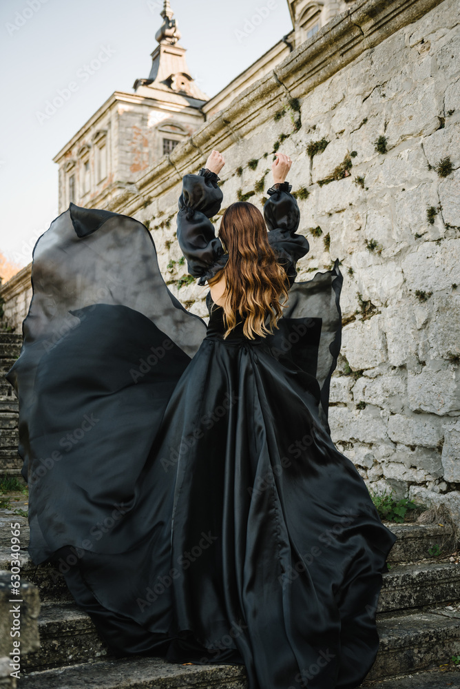 Woman in long black dress stands on stairs near ancient palace at sunset. Model girl in nature autumn day. Luxury female near old Pidhirtsi Castle, Lviv region, Ukraine. Stylish bride. Back view. Woman in long black dress stands on stairs near ancient palace at sunset. Model girl in nature autumn day. Luxury female near old Pidhirtsi Castle, Lviv region, Ukraine. Stylish bride. Back view.