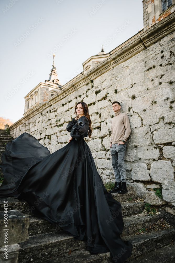 Woman in black long dress with man stand on stairs near ancient palace at sunset. Female, male walk near brick walls in street. Luxury couple newlyweds near old Castle. Stylish bride and groom outdoor